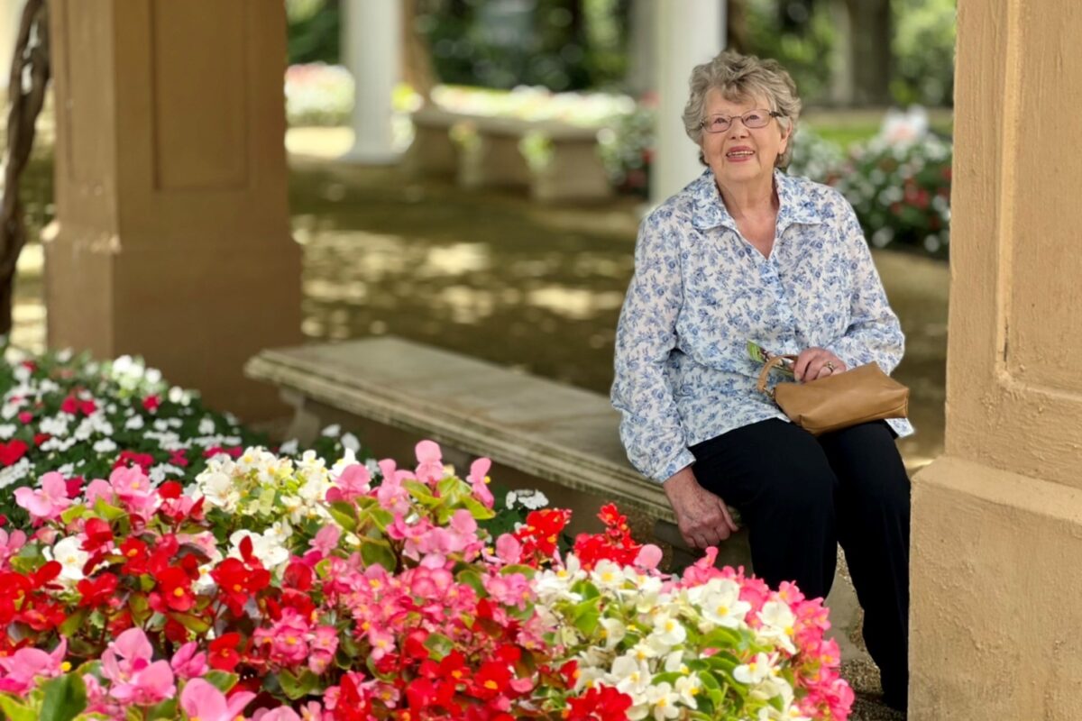 Eleanor sits on a park bench, in the foreground are a mass of colourful pink, red and white petunias.